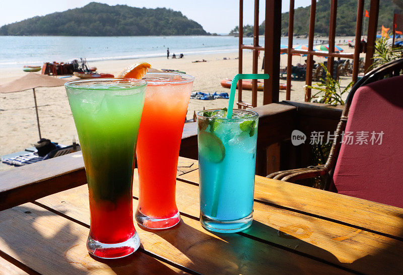 在露天海滩酒吧餐厅的木桌上的一排两种鸡尾酒和无酒精鸡尾酒的图像，彩虹模拟鸡尾酒与橙色片装饰，龙舌兰日出，蓝色泻湖伏特加和蓝色cura<s:1>鸡尾酒，吸管，沙子和大海的背景